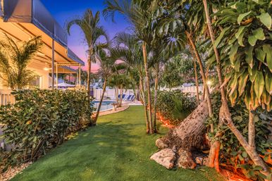 Tropical backyard with manicured lawn and palm trees leading to a swimming pool and lounge chairs, warm outdoor lighting and colorful sunset sky