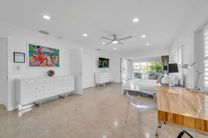 Bright modern coastal master bedroom with glossy tile floors, white dressers and bed, wooden desk with clear acrylic chair, ceiling fan, colorful tropical painting, and sliding glass doors opening to a balcony overlooking palm trees and waterfront.