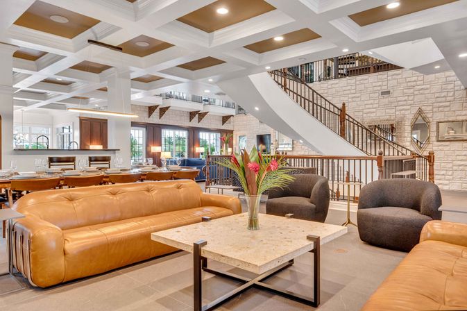 Bright luxury hotel lobby with tan leather sofas, rounded gray lounge chairs and a stone-topped coffee table with tropical flowers, grand curved staircase, stone accent wall, coffered ceiling and adjacent dining area.