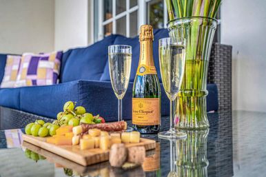 Cozy patio scene: bottle of champagne and two filled flutes beside a cheese-and-grape board on a glass table, with a navy outdoor sofa, patterned pillows, and a tall vase of green stems in the background.