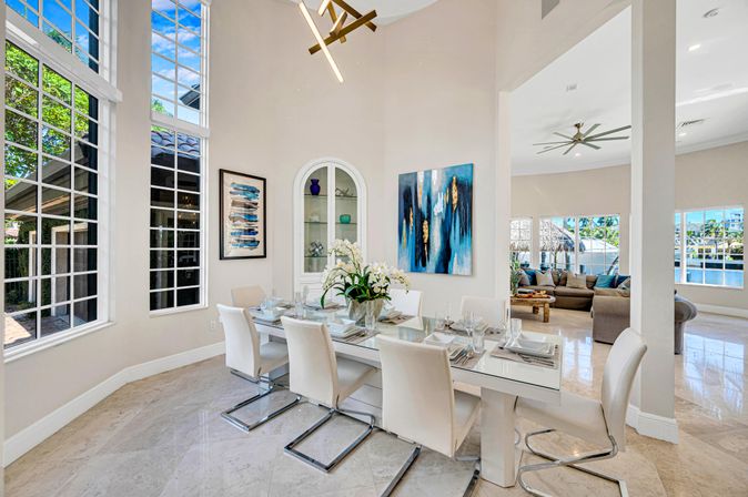 Sunlit luxury dining room with high ceilings and floor-to-ceiling windows overlooking a waterfront canal, glass dining table set with white leather chairs, marble floors, modern chandelier and blue abstract wall art