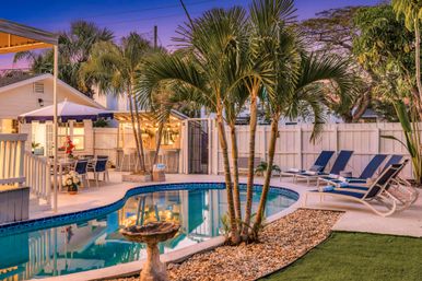 Tropical backyard pool at twilight with palm trees, curved blue-tiled swimming pool, navy lounge chairs, outdoor dining under an umbrella and warm patio lights