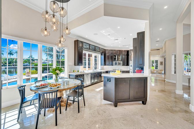 Sunlit luxury waterfront open-plan kitchen with dark wood cabinets, marble floors, breakfast island and round dining table under glass pendant lights, large windows overlooking pool, canal and palm trees.