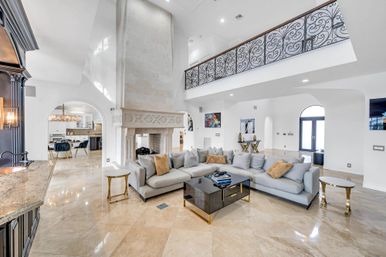 Airy luxury open-plan living room with high ceilings and marble floors, oversized carved stone fireplace, light gray sectional sofa, black glass coffee table with gold accents, and decorative wrought-iron mezzanine railing — bright Mediterranean-style interior.