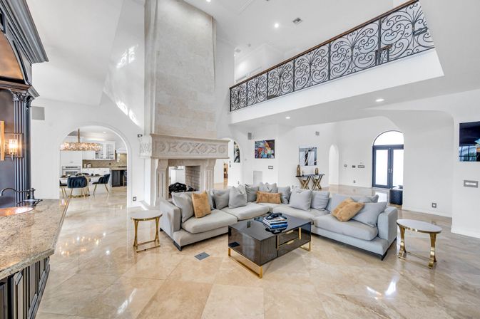 Airy luxury open-plan living room with high ceilings and marble floors, oversized carved stone fireplace, light gray sectional sofa, black glass coffee table with gold accents, and decorative wrought-iron mezzanine railing — bright Mediterranean-style interior.