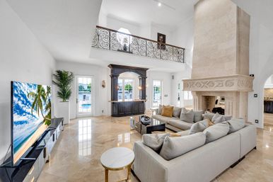 Sunlit coastal luxury living room with double-height ceiling, marble floors, oversized carved stone fireplace, large neutral sectional, dark wood wet bar, ornate wrought-iron balcony and waterfront views.