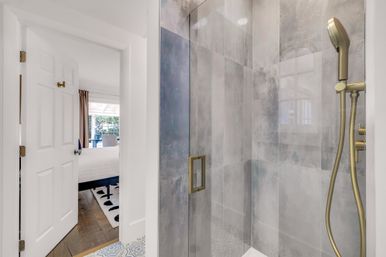 Sleek modern ensuite with glass shower enclosure, gray stone-look tile walls and brass handheld showerhead, open door revealing a bright bedroom with bed and window.