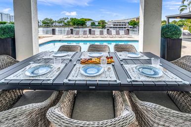 Poolside waterfront patio with a dark slatted dining table set for six—blue ceramic plates, crystal glasses, charcuterie board, woven wicker chairs, pool and calm bay view with lounge chairs.