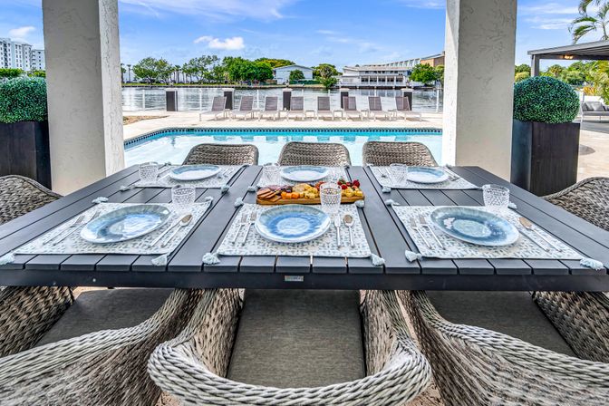 Poolside waterfront patio with a dark slatted dining table set for six—blue ceramic plates, crystal glasses, charcuterie board, woven wicker chairs, pool and calm bay view with lounge chairs.