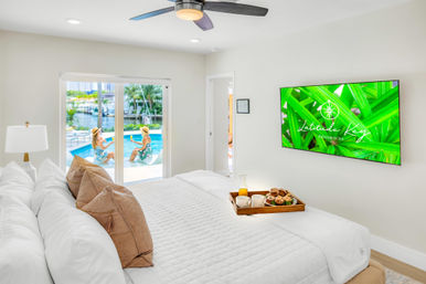 Bright waterfront resort bedroom with white quilted bed, tan pillows, breakfast tray, ceiling fan and wall TV with tropical graphic; sliding glass doors open to pool and marina with palm trees
