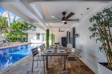 Covered tropical backyard patio with a long outdoor dining table set for eight, modern ceiling fans and tiled floor beside a blue swimming pool and lush palm landscaping.