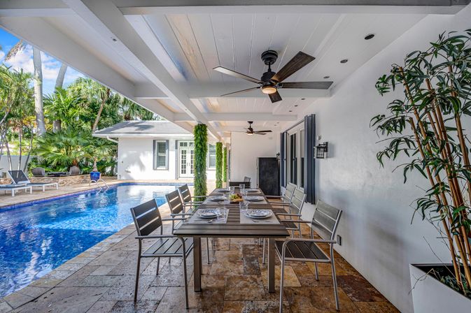 Covered tropical backyard patio with a long outdoor dining table set for eight, modern ceiling fans and tiled floor beside a blue swimming pool and lush palm landscaping.
