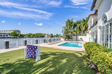 Sunny waterfront pool patio at a luxury home along a canal with palm trees, private dock, loungers, cabana and an oversized outdoor Connect Four on the lawn.
