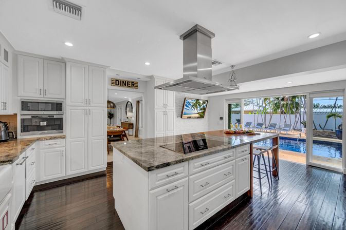 Bright modern kitchen with marble-topped island and stainless range hood, white cabinetry and dark hardwood floors, sliding glass doors opening to a pool and palm‑lined patio.