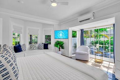 Sunlit coastal-style master bedroom with crisp white bedding, window seat with navy accent pillows, wall-mounted TV, potted palm, and sliding glass doors opening to a tropical garden patio.