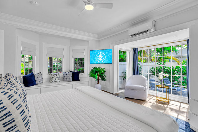 Sunlit coastal-style master bedroom with crisp white bedding, window seat with navy accent pillows, wall-mounted TV, potted palm, and sliding glass doors opening to a tropical garden patio.