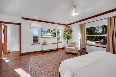Sunlit tropical coastal bedroom with hardwood floors, white bed and armchair, ceiling fan, wall TV, and large window framing palm trees and a beach view.