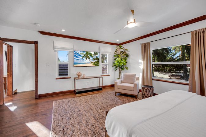 Sunlit tropical coastal bedroom with hardwood floors, white bed and armchair, ceiling fan, wall TV, and large window framing palm trees and a beach view.