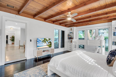 Sunny coastal-style bedroom with exposed wood-beam ceiling, white bedding and bunk bed, TV console, built-in bookshelves and large windows overlooking greenery.