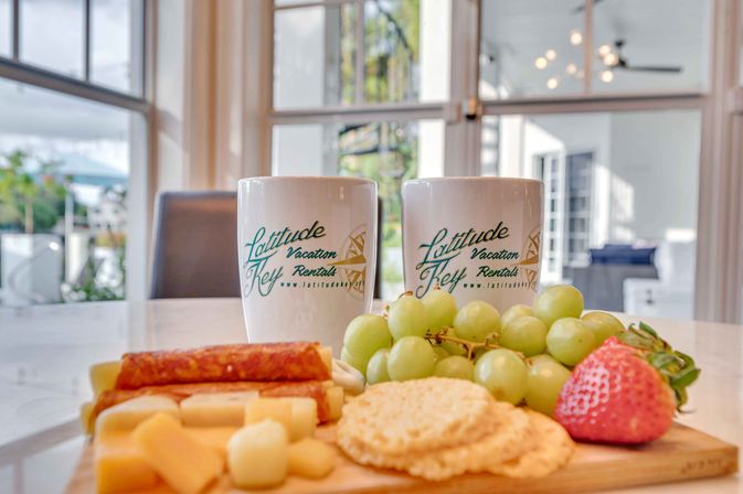 Bright coastal vacation-rental kitchen: two white branded coffee mugs behind a cheerful breakfast charcuterie board with green grapes, a ripe strawberry, cheese, crackers and sliced sausage.