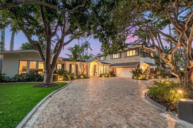 Twilight view of a luxury two-story home with a wide brick-paver driveway, glowing entry, attached garage, mature trees and tropical landscaping with palm trees.