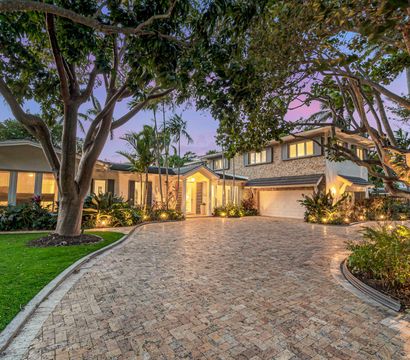 Twilight view of a luxury two-story home with a wide brick-paver driveway, glowing entry, attached garage, mature trees and tropical landscaping with palm trees.