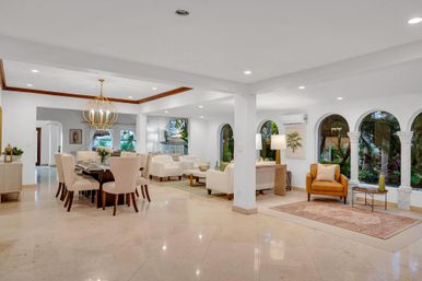 Open-concept living and dining room with polished marble floors, neutral upholstered seating, a gold chandelier over a dark wood table, Mediterranean arched windows framing lush tropical garden views and a cozy tan leather armchair.