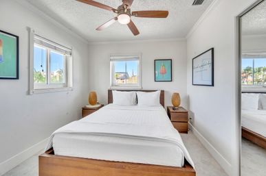 Bright coastal-style bedroom with white linens on a wooden platform bed, matching wood nightstands with woven lamps, ceiling fan, two windows with palm-tree views, and playful popsicle wall art.