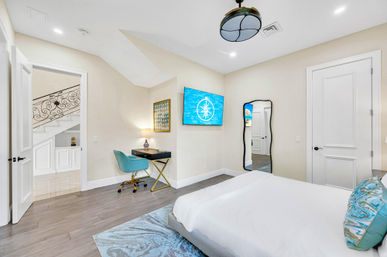 Bright modern bedroom with coastal accents — white bed with turquoise pillows, hardwood floors, wall-mounted TV with compass graphic, gold-legged desk and teal swivel chair, full-length mirror and view to staircase landing.
