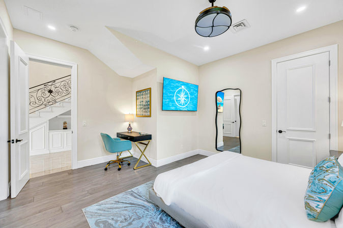 Bright modern bedroom with coastal accents — white bed with turquoise pillows, hardwood floors, wall-mounted TV with compass graphic, gold-legged desk and teal swivel chair, full-length mirror and view to staircase landing.