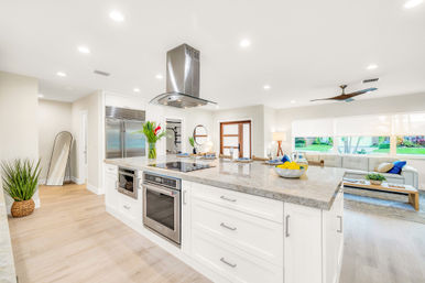 Sunlit open-concept kitchen and living area featuring a large marble island with cooktop and stainless-steel hood, built-in oven and refrigerator, white cabinetry, light wood floors, potted plants, and a cozy sofa by a wide window with garden views.