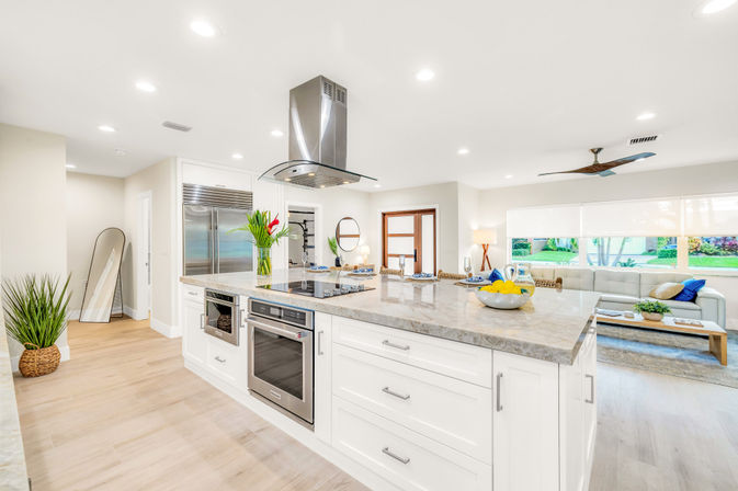 Sunlit open-concept kitchen and living area featuring a large marble island with cooktop and stainless-steel hood, built-in oven and refrigerator, white cabinetry, light wood floors, potted plants, and a cozy sofa by a wide window with garden views.