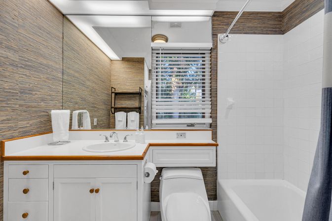 Bright coastal-style bathroom with white vanity and wood trim, large mirror, toilet, tiled bathtub with shower, and window blinds showing palm trees