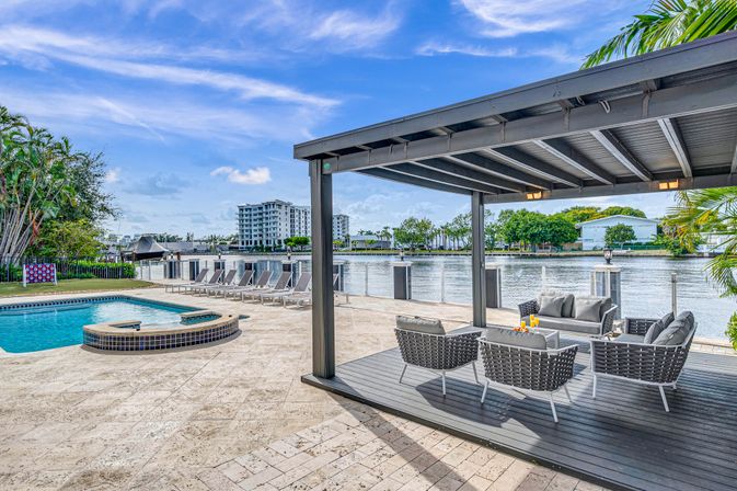 Poolside waterfront patio with raised spa, row of sun loungers and a covered pergola seating area with wicker sofas overlooking a calm canal, palm trees and distant waterfront condos under a blue sky.