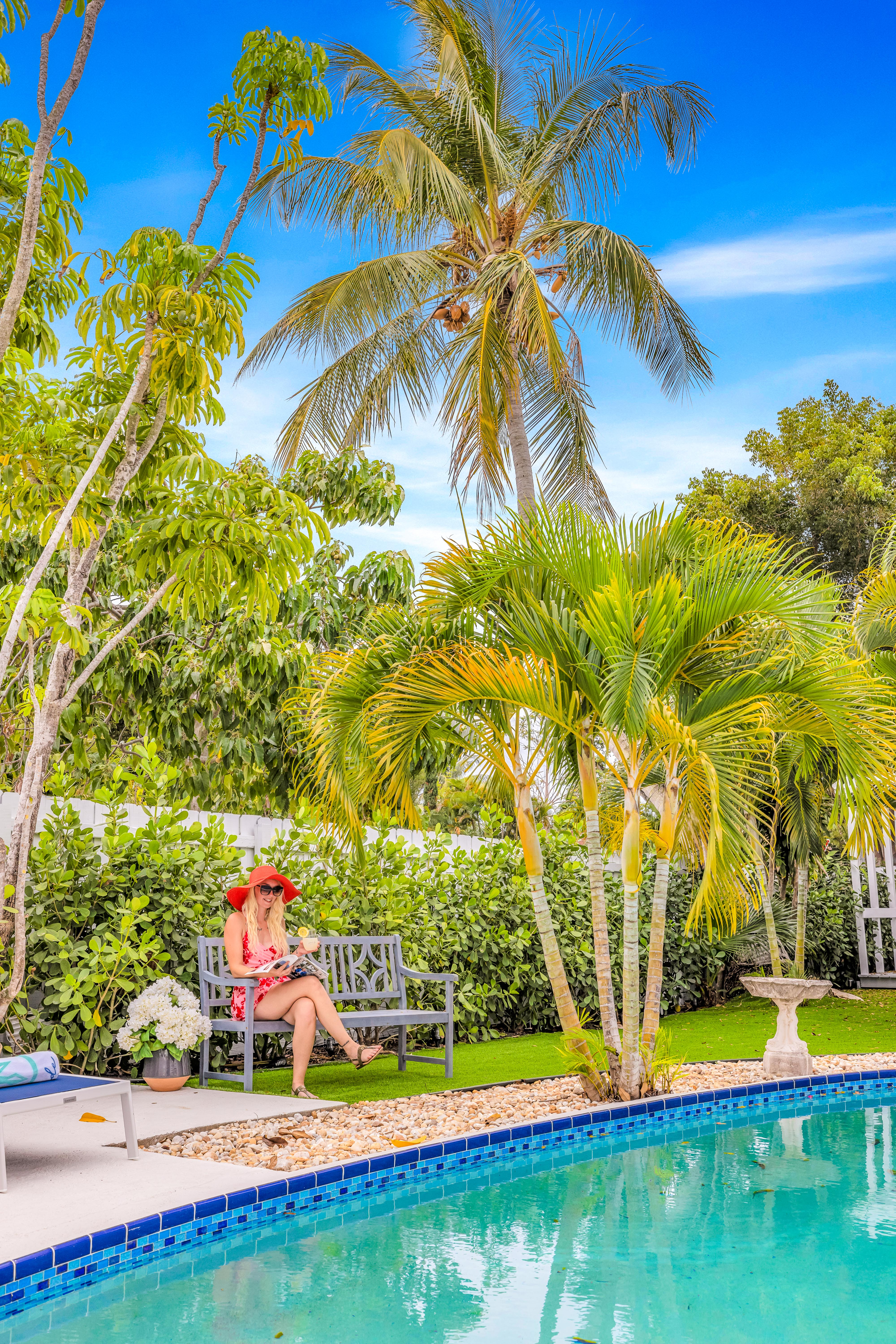 Tropical backyard poolside: woman in a red sunhat reads on a bench beside a turquoise swimming pool, framed by palm trees, lush hedges and a bright blue sky.