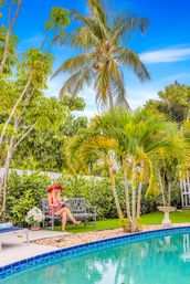 Tropical backyard poolside: woman in a red sunhat reads on a bench beside a turquoise swimming pool, framed by palm trees, lush hedges and a bright blue sky.