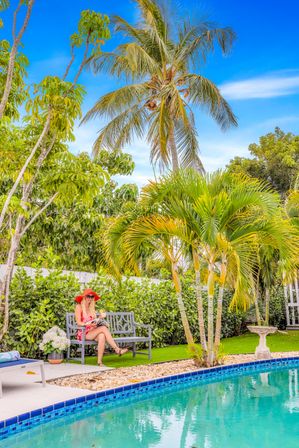 Tropical backyard poolside: woman in a red sunhat reads on a bench beside a turquoise swimming pool, framed by palm trees, lush hedges and a bright blue sky.