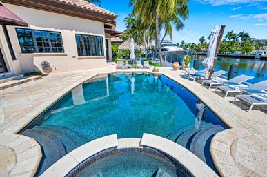 Sunlit tropical waterfront villa with a curved infinity-style pool and attached spa, stone terrace with lounge chairs and umbrellas, palm trees, and a private dock with a yacht on a calm canal.