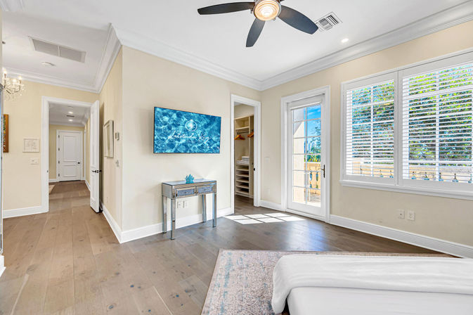 Sunlit master bedroom with hardwood floors, ceiling fan, wall-mounted TV, mirrored console, walk-in closet and French doors opening to a balcony with plantation-shuttered windows.