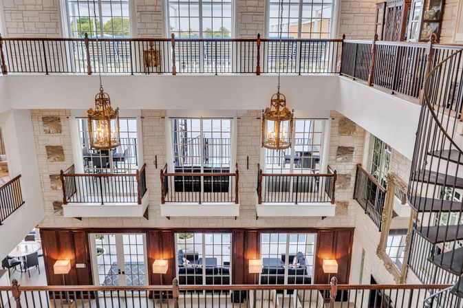 Sunlit multi-level atrium with stone walls, iron balcony railings, ornate hanging chandeliers, large windows, spiral staircase and a ground-floor seating area.