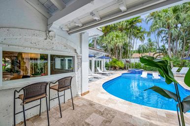 Sunny tropical backyard with a curved blue swimming pool and raised spa, travertine tile patio, two wicker bar stools under a covered porch, white lounge chairs and umbrellas, and tall palm trees