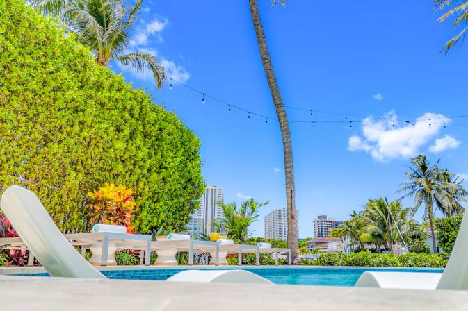 Sunny tropical poolside with white lounge chairs, palm trees, string lights and a distant coastal city skyline under a blue sky