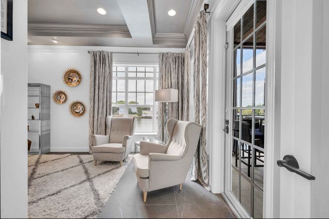 Sunlit waterfront living room with two beige wingback chairs by a large window overlooking a lake, patterned curtains, shag rug and glass patio door.
