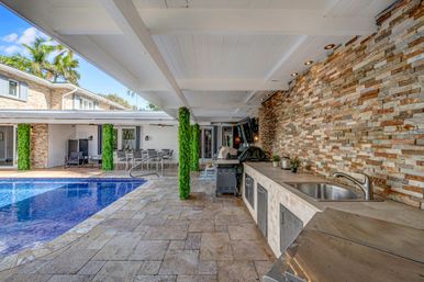Resort-style covered patio with blue swimming pool, built-in outdoor kitchen and grill, stone veneer accent wall, dining area and palm trees