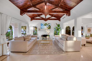 Sunlit tropical coastal villa living room with a dramatic dark wood vaulted ceiling, two beige sofas facing a white marble fireplace and glass coffee table, arched openings and large windows framing palm trees and garden views.