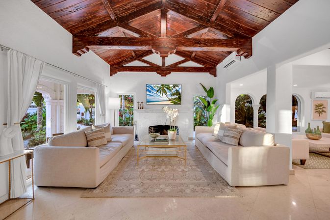 Sunlit tropical coastal villa living room with a dramatic dark wood vaulted ceiling, two beige sofas facing a white marble fireplace and glass coffee table, arched openings and large windows framing palm trees and garden views.