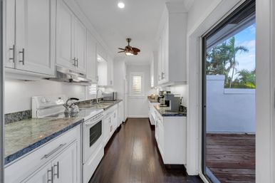 Bright white galley kitchen with granite countertops, dark hardwood floors, white stove and microwave, ceiling fan and coffee station, sliding glass door to patio with palm trees.