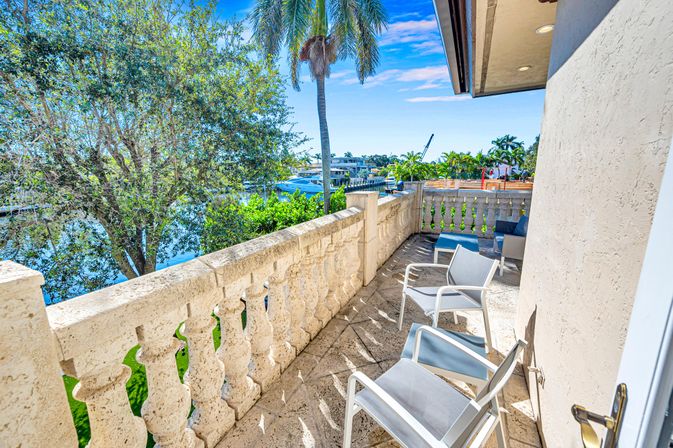 Sunny tropical waterfront balcony with stone balustrade and white patio chairs overlooking a palm‑fringed canal marina with boats