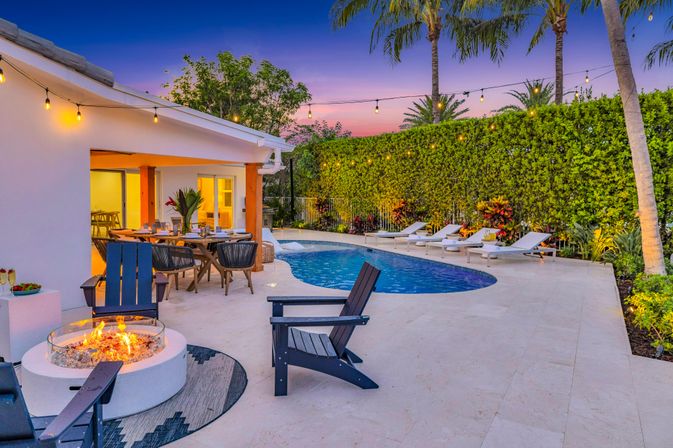 Sunset tropical backyard patio with palm trees and string lights, curved blue swimming pool, row of white loungers, outdoor dining table and a glowing fire pit on a tiled deck.