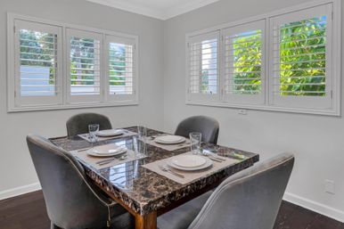 Modern dining nook with dark marble table set for four, grey upholstered chairs, and white plantation shutters revealing a leafy backyard view